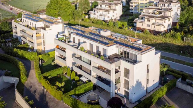 Luftaufnahme mehrerer moderner Mehrfamilienhäuser. Auf den Dächern sind Photovoltaikanlagen installiert, mit denen die Elektroautos in den Tiefgaragen mit sauberem Strom geladen werden.