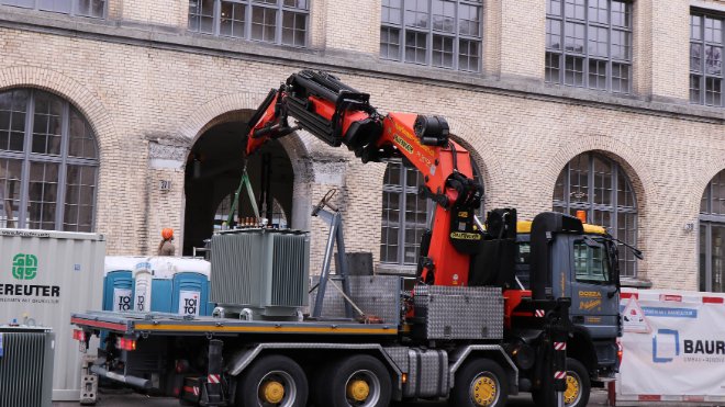 Der Transformator wird vorsichtig mit dem Kran vom Laster gehoben. Mit dem Ausleger wird der Trafo in die schmale Öffnung manövriert. 